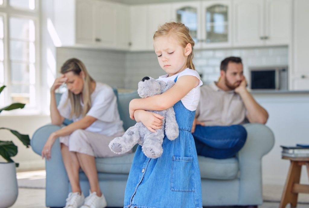 Young girl holding a teddy bear while her parents sit apart in the background, illustrating the emotional impact of divorce on children.