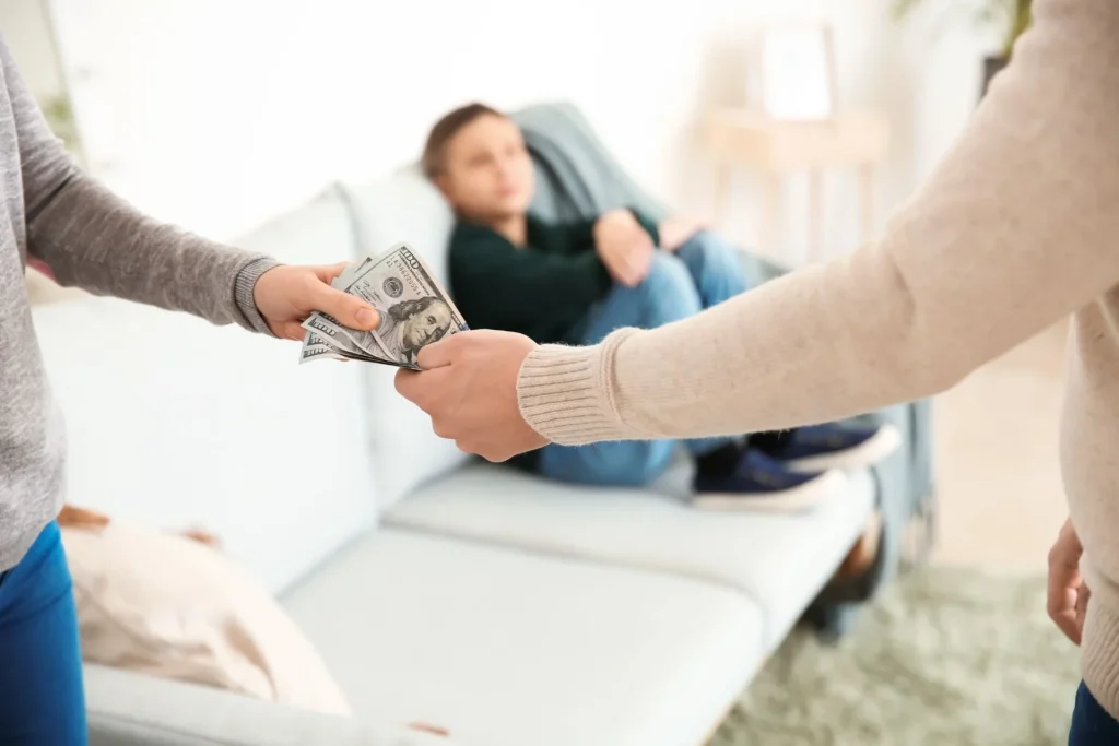 Parent handing money to another parent while a child sits in the background, illustrating child support or family financial assistance.