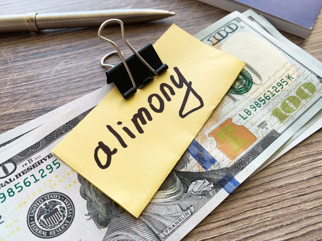 Sticky note labeled 'alimony' clipped to U.S. dollar bills on a desk, representing spousal support payment.